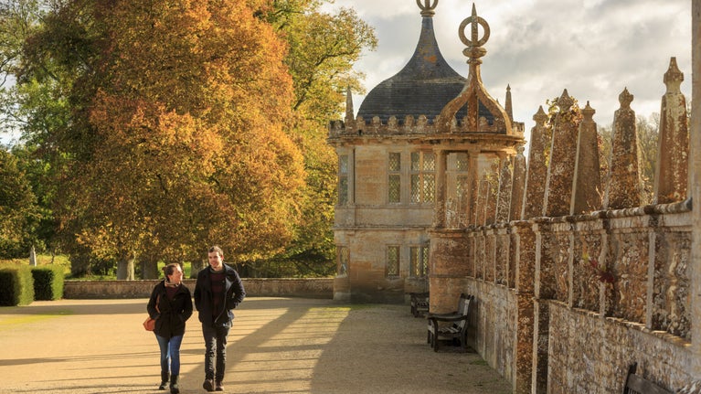 Two visitors walk past ornate walls in a garden, with trees in autumn leaf behind them